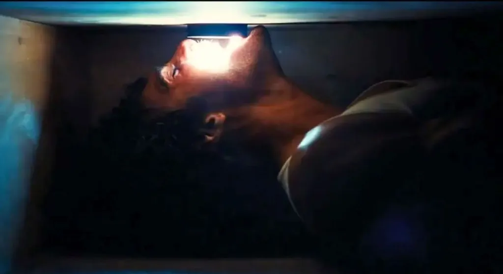 Jacob Elordi lying on his back in a dark space, illuminated by a bright light above his head.