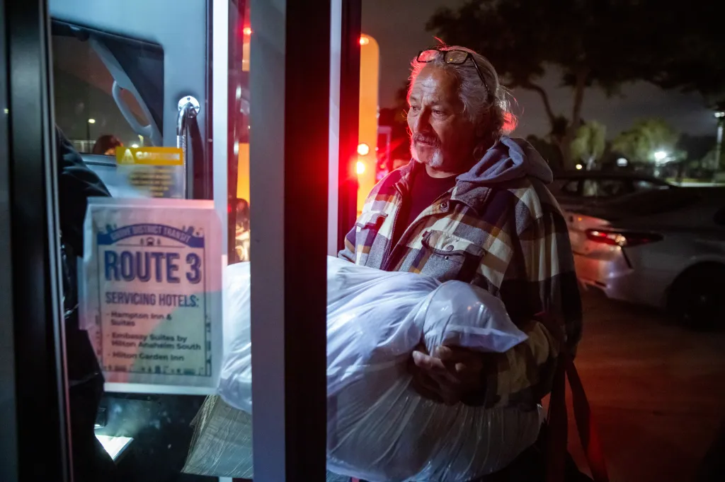 A man carrying belongings wrapped in plastic walks from an aerospace chemical plant evacuee shelter to a bus for new shelter.