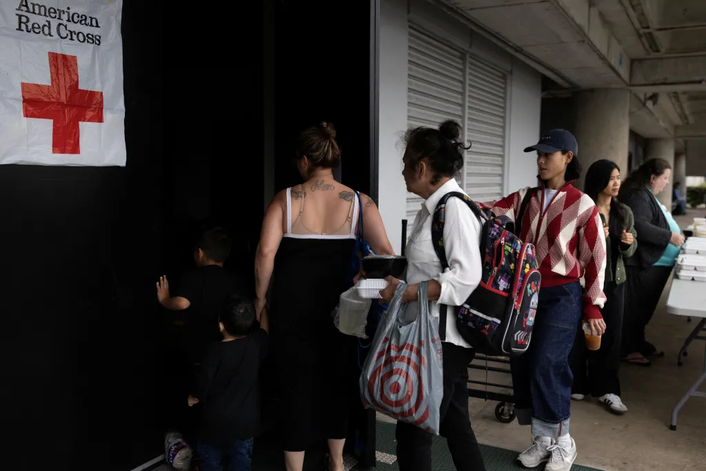 Evacuees enter a shelter with a red cross sign, carrying food containers and bags.