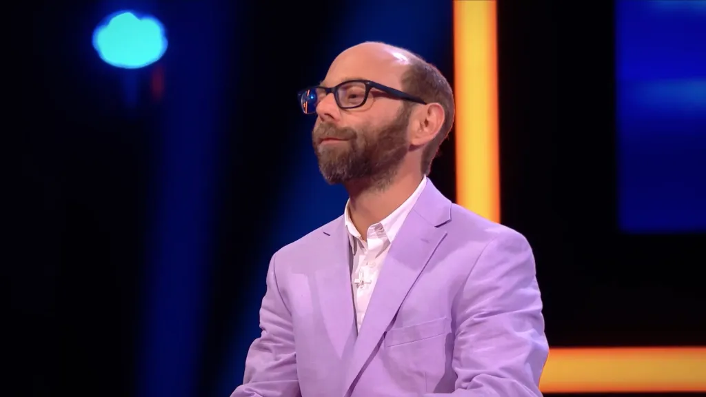 Steven Anthony Lawrence, known for playing Beans on Even Stevens, wearing a light purple blazer and white shirt with a cross necklace on a game show set.