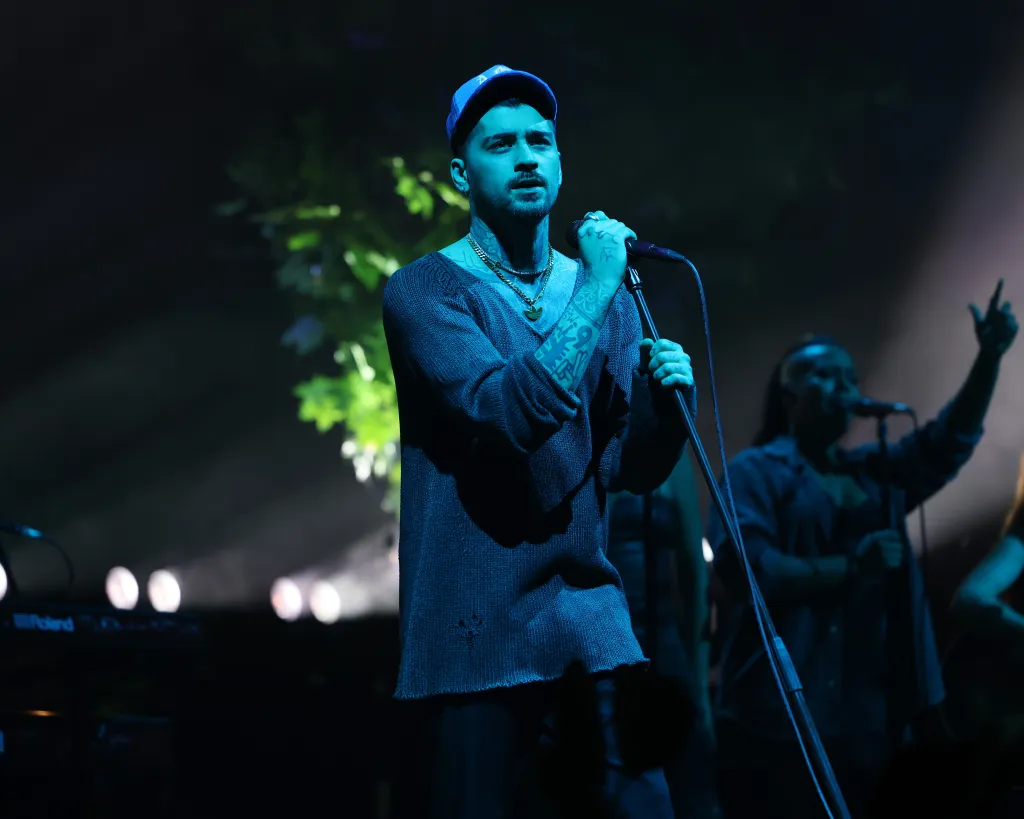 Zayn Malik performing on stage in a blue cap, holding a microphone.
