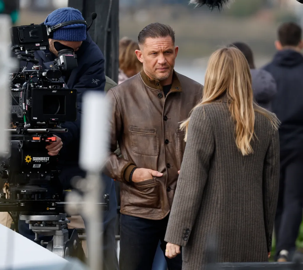 Tom Hardy on the set of MobLand, wearing a brown leather jacket and looking at an unseen woman.