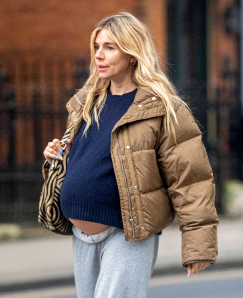 Sienna Miller, heavily pregnant, walks down the street in a brown puffer jacket, navy sweater, and grey sweatpants with a zebra print shoulder bag.