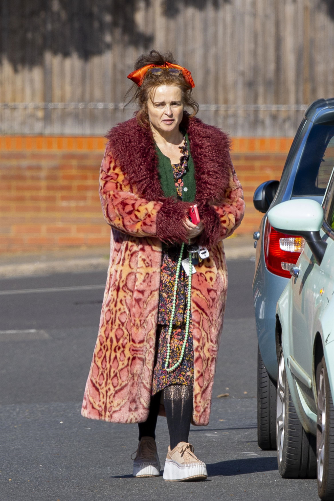 Helena Bonham-Carter in a long patterned coat with fur trim, floral dress, green cardigan, black tights, and platform shoes. She wears a red headscarf, sunglasses on her head, and a beaded necklace. She is holding keys and a phone.