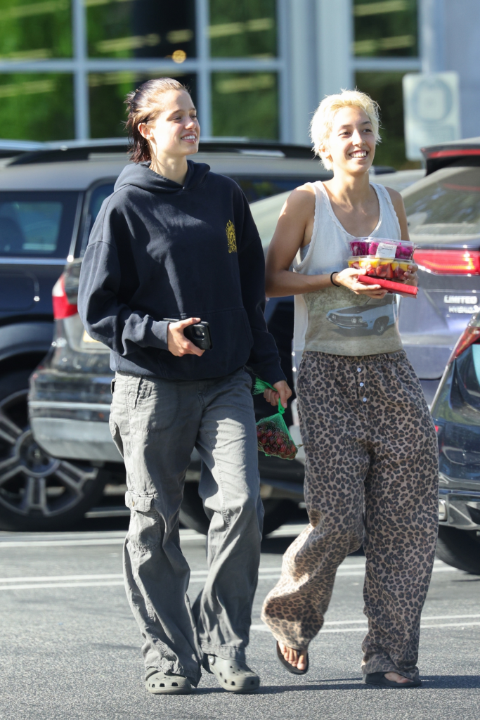 Shiloh Jolie-Pitt in a blue hoodie and gray cargo pants walking next to Keoni Rose in an animal print tank top and pants, carrying fruit.