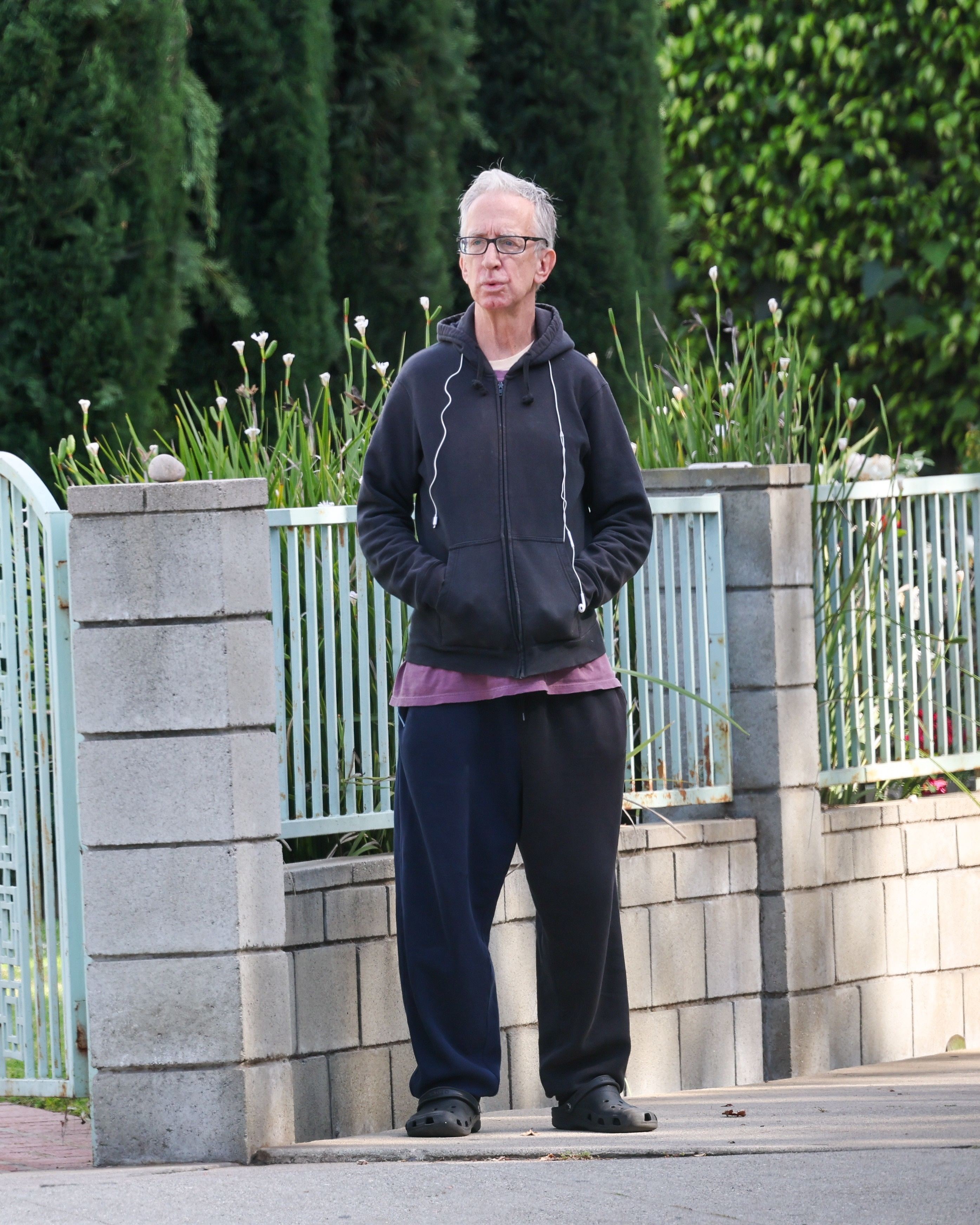 Andy Dick in a black hoodie, dark sweatpants, and black Crocs, standing on a street next to a fence.