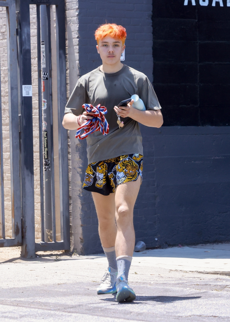 Knox Jolie-Pitt with bright orange hair, wearing a gray t-shirt and tiger-print shorts, holding a phone and water bottle.