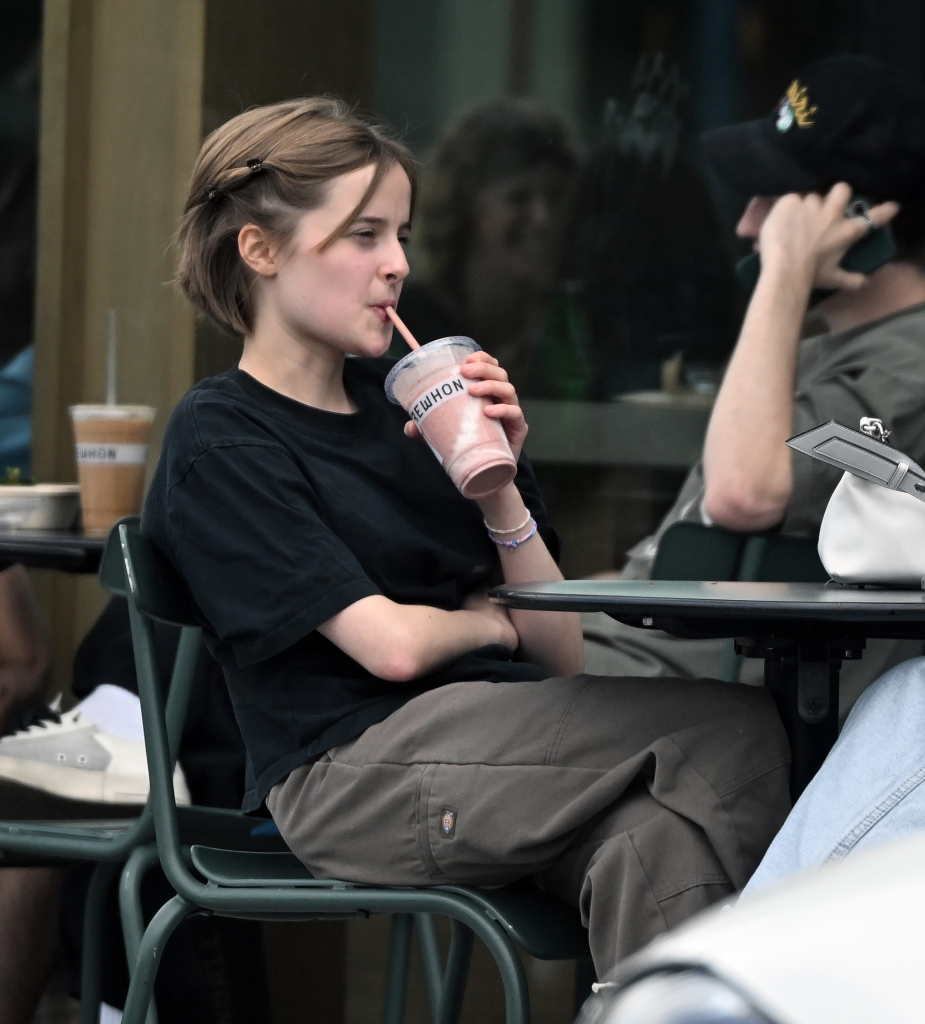 Vivienne Jolie, in a black T-shirt and grey pants, sitting outside Erewhon and drinking a Hailey Bieber smoothie.