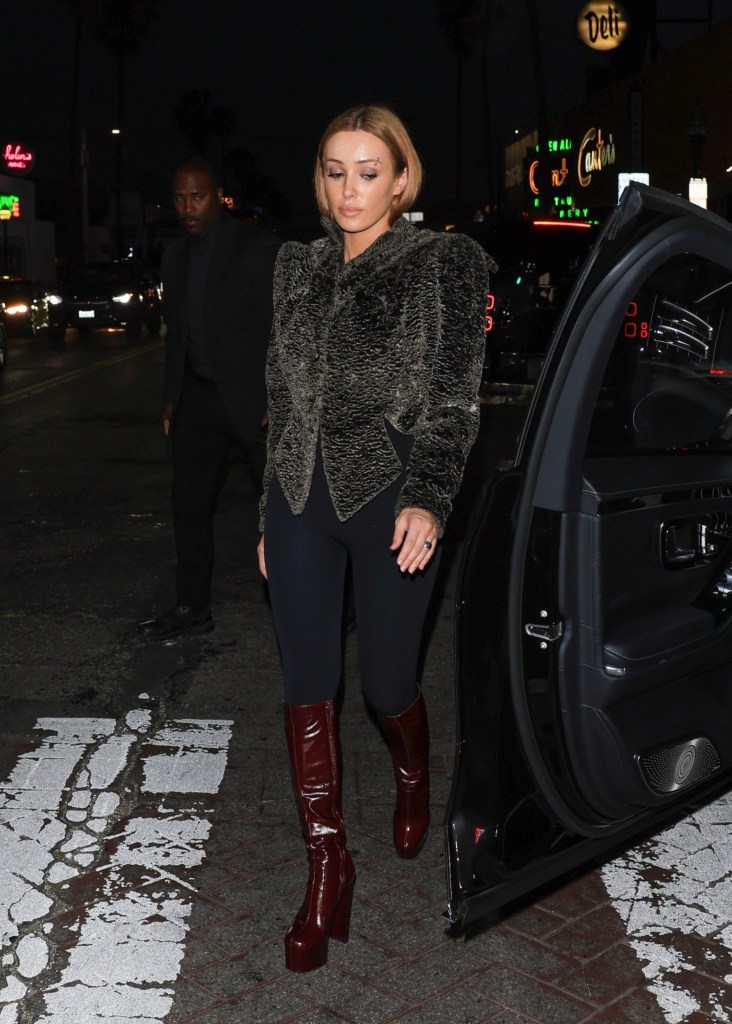 Bianca Censori steps out of a car at night in a dark, textured jacket, black leggings, and high burgundy platform boots.