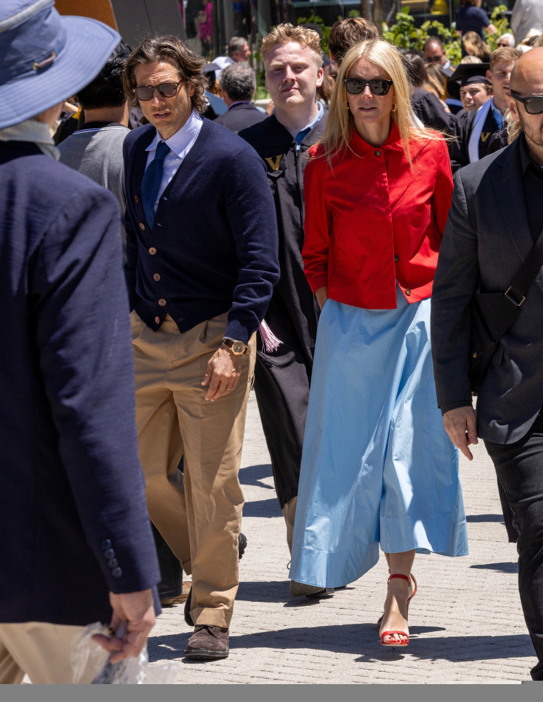Gwyneth Paltrow in a red jacket and light blue maxi skirt, walking next to Brad Falchuk, wearing a navy cardigan over a light blue shirt and tan trousers.