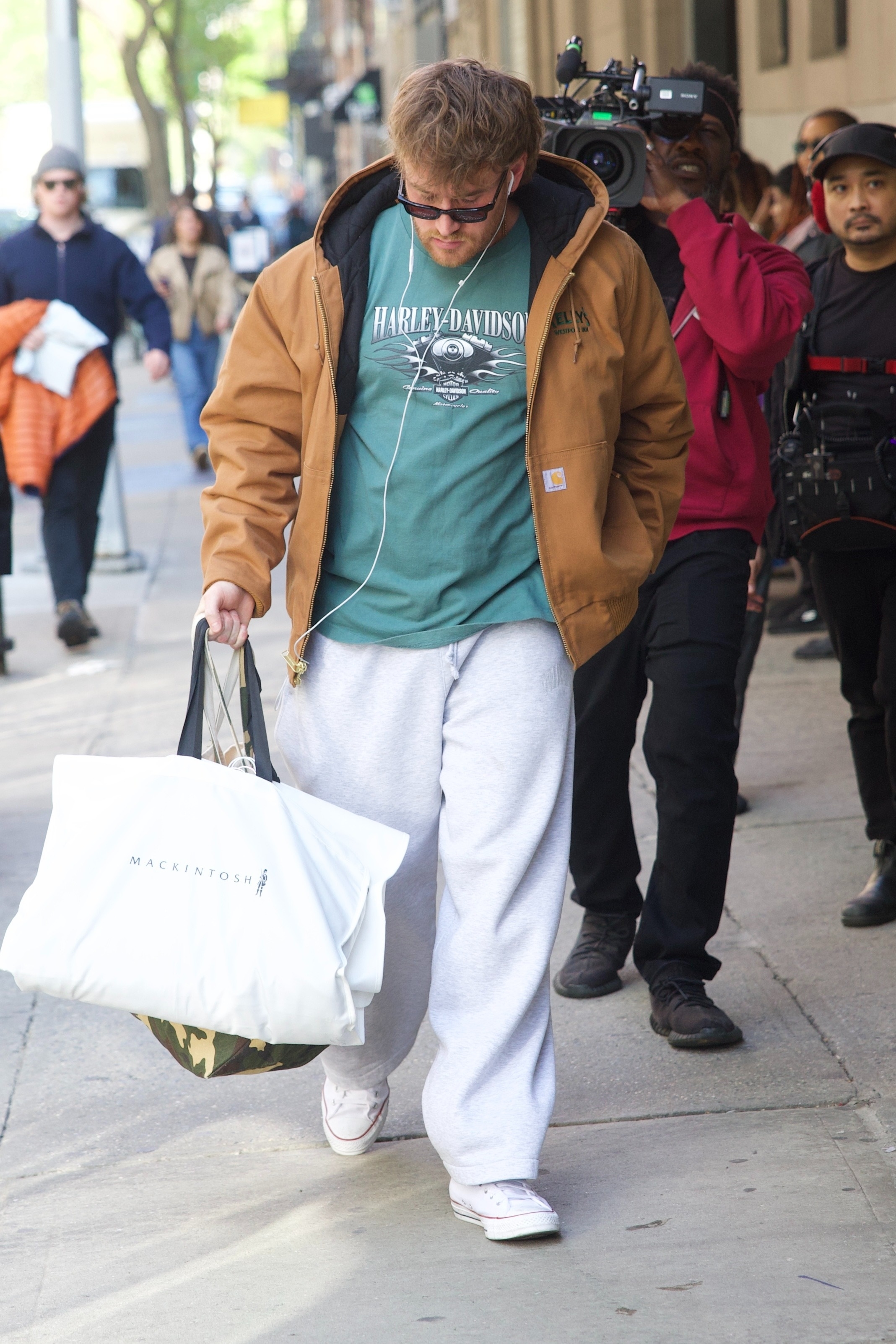 West Wilson from Summer House walking on a city street carrying a Mackintosh shopping bag, with a camera crew behind him.