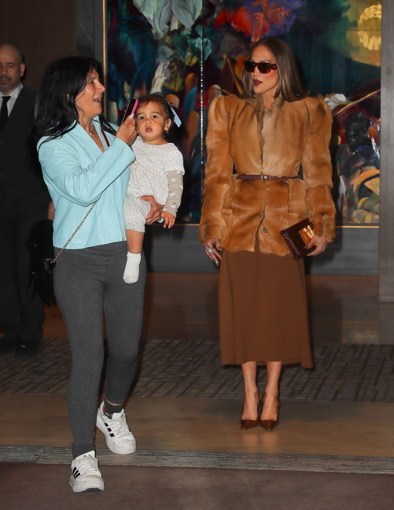 Jennifer Lopez posing for photos outside a New York City hotel, while a woman holding a baby walks in front of the cameras.