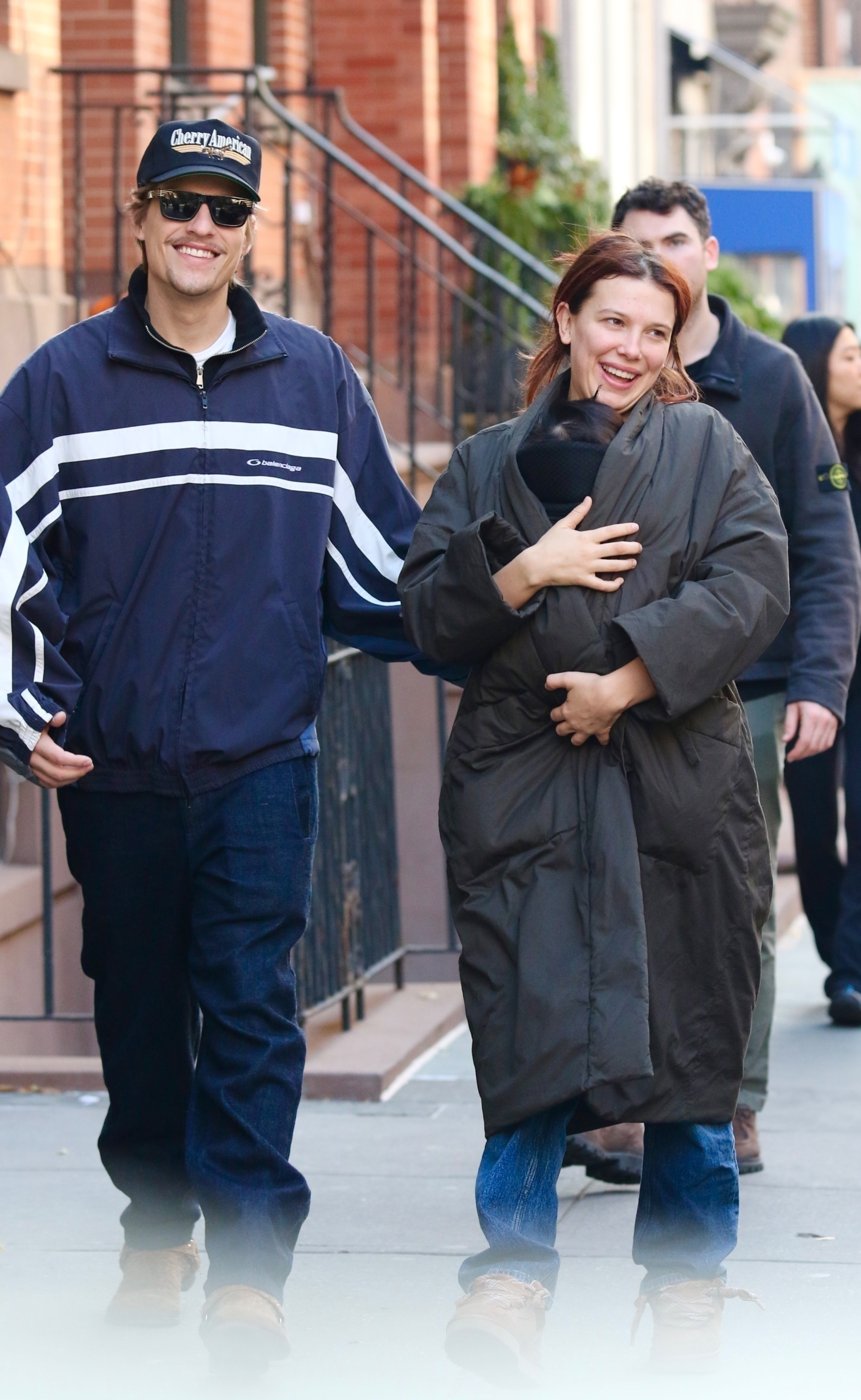 Millie Bobby Brown smiling while holding her baby in a carrier and Jake Bongiovi smiling beside her.