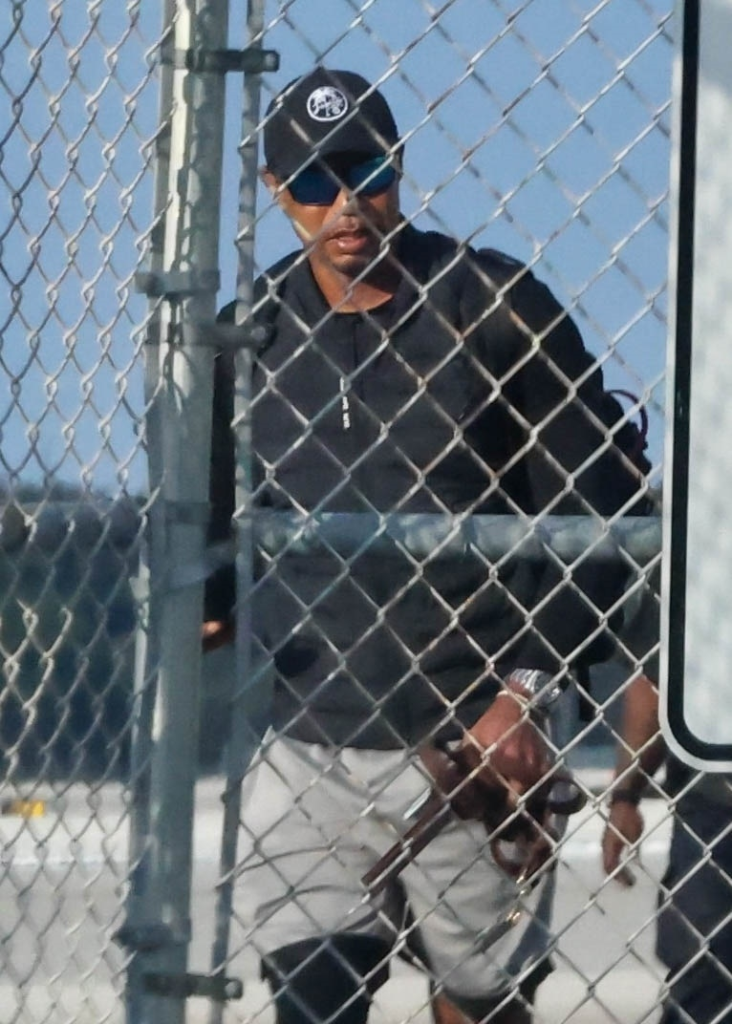 Tiger Woods wearing a black cap, sunglasses, and a black jacket behind a chain-link fence.