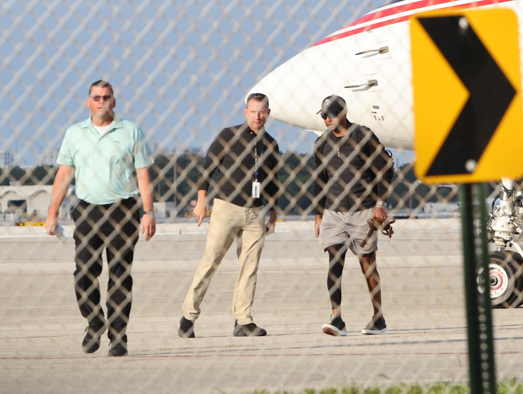 Golfer Tiger Woods, in a baseball cap and sunglasses, walks on an airfield with two other men, a private jet and a yellow directional sign in the background.