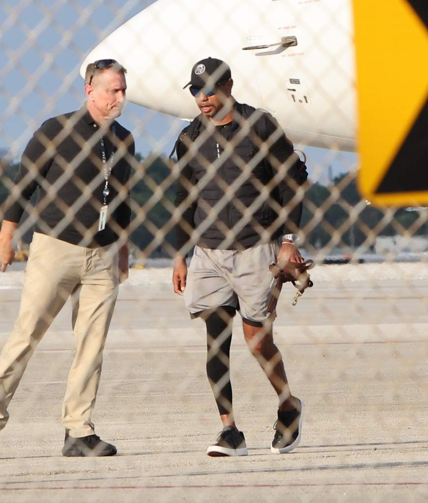 Golfer Tiger Woods returns home after rehab, walking with a dog leash in hand, accompanied by another man, near a private jet.