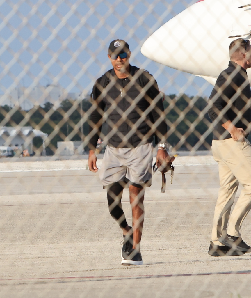 Golfer Tiger Woods, wearing sunglasses and a baseball cap, walks on an airfield with a dog leash in his hand.