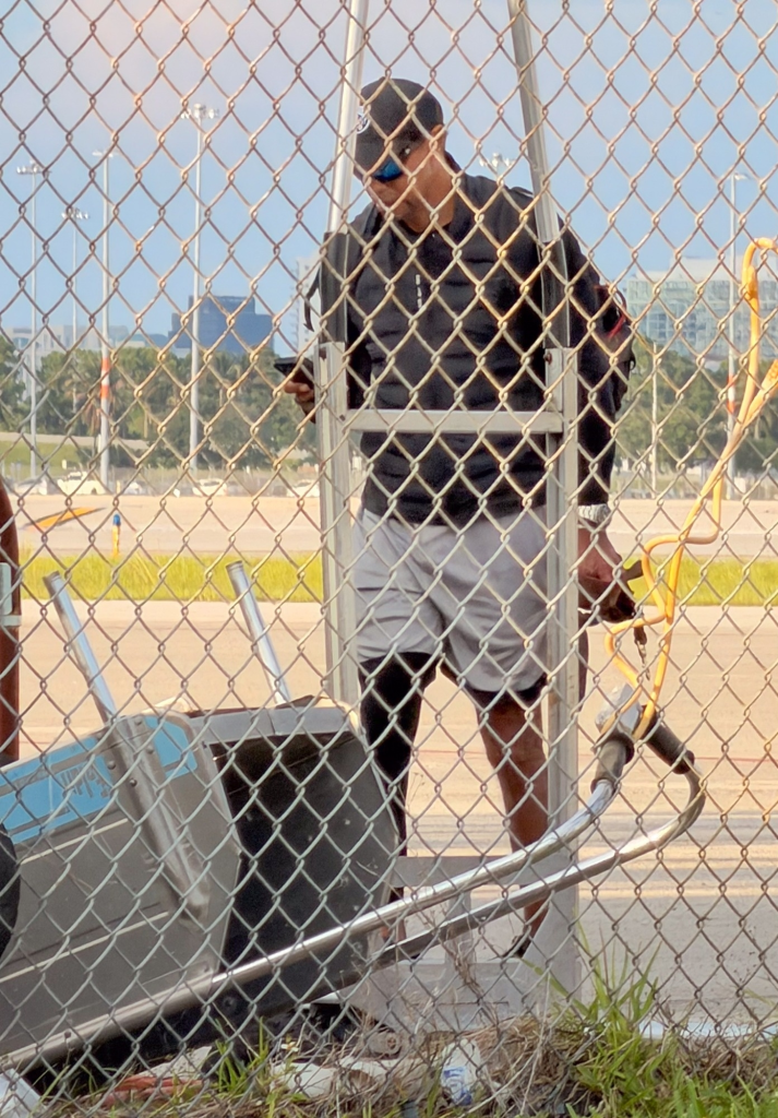 Tiger Woods wearing a black jacket, grey shorts, baseball cap, and blue sunglasses behind a chain-link fence.