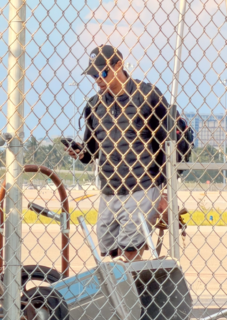 Tiger Woods at an airport, seen through a chain-link fence, wearing sunglasses, a black baseball cap, black jacket, and gray shorts, holding a phone.