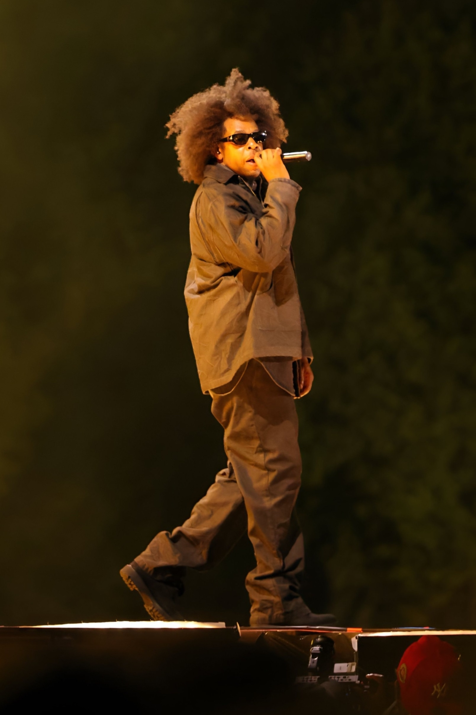 Jay-Z, wearing sunglasses and a dark outfit, performs on stage with natural, bushy hair while holding a microphone.