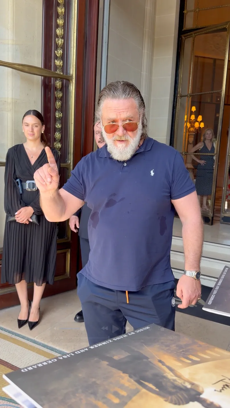 Russell Crowe, in a navy polo shirt and sunglasses, holds up a finger while talking to autograph seekers.