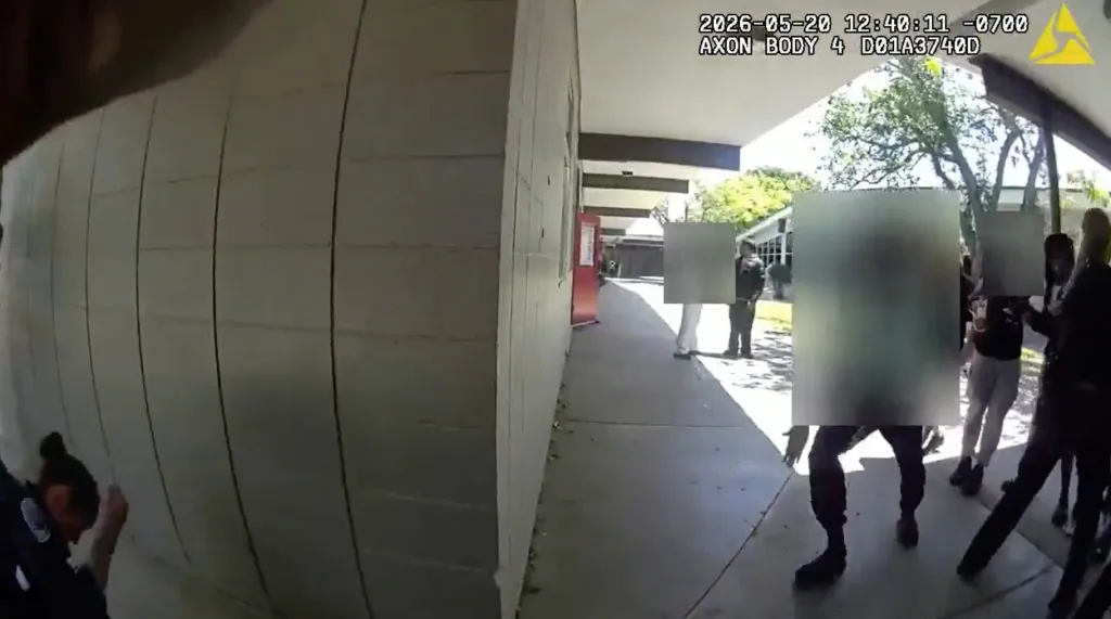 Still from a bodycam video showing a police officer with a student against a wall while other students and an officer are blurred in the background.