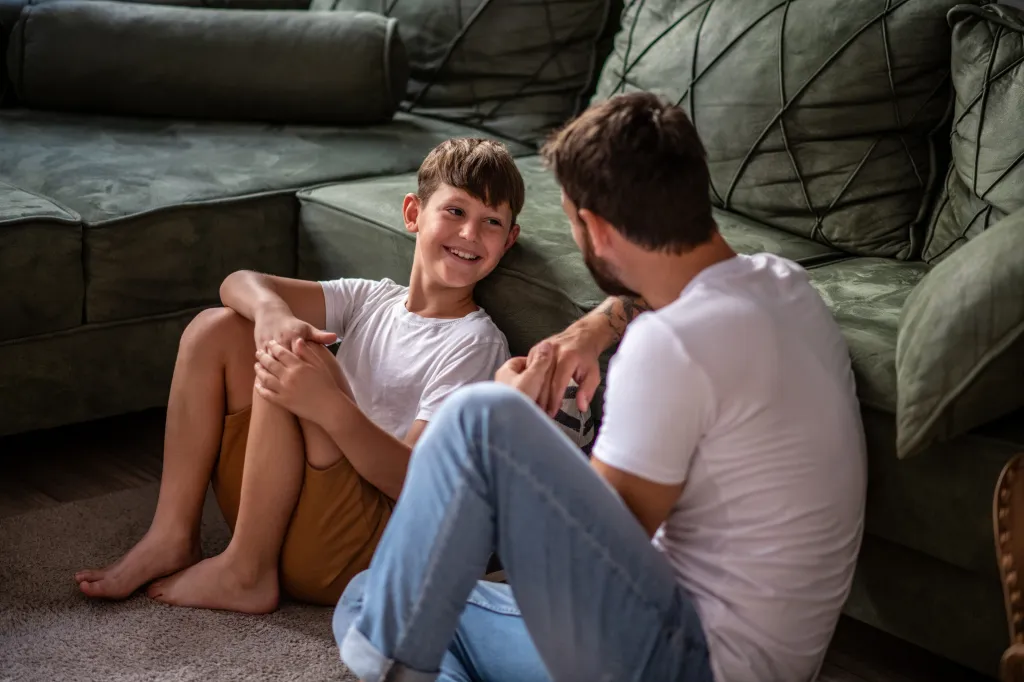 Father and son sitting on the living room floor and talking.