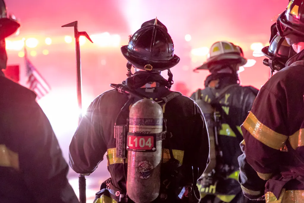 Firefighters with breathing apparatus and an American flag in the background at the scene of a 4-alarm fire in Brooklyn.