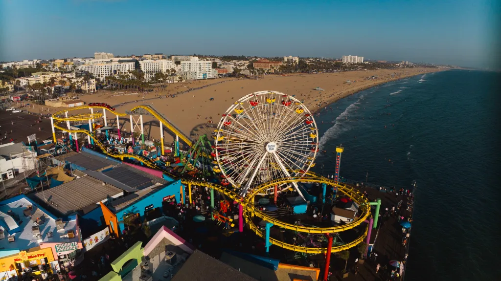Drone view of the Santa Monica Pier, showing a ferris wheel, rollercoaster, and the beach with city buildings in the background.