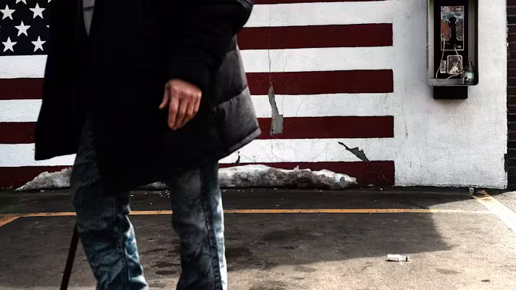 A person, visible torso down, walks through an economically stressed urban area, with a mural of an American flag behind them.