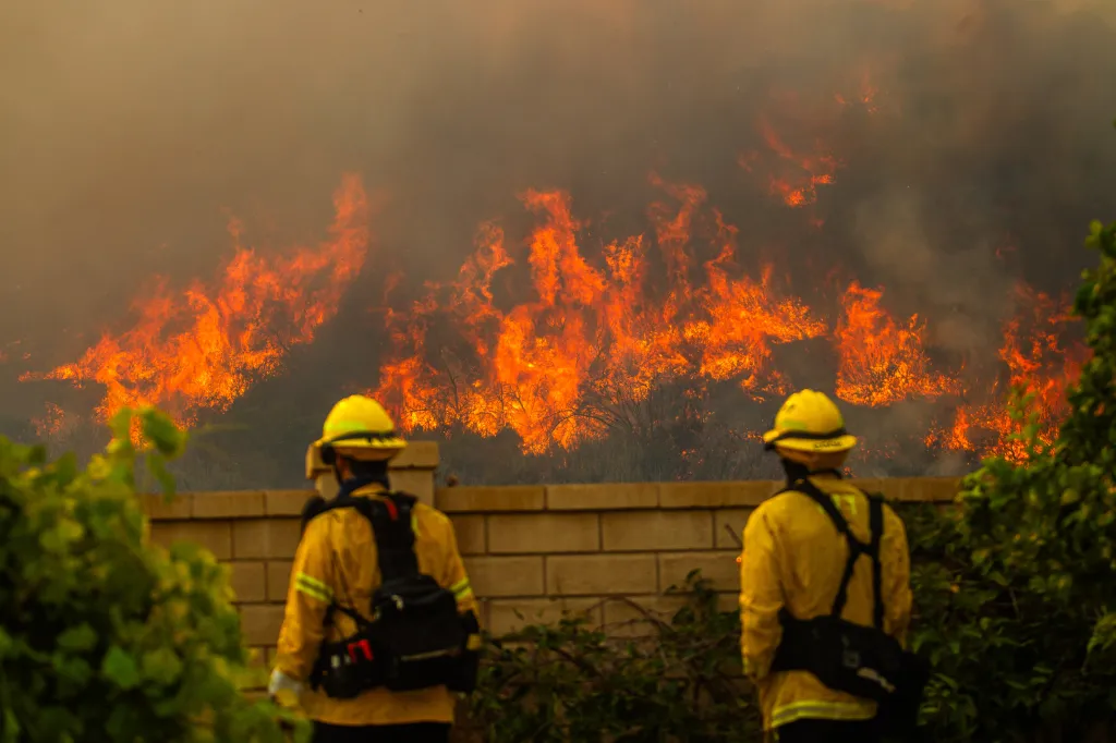 Two firefighters monitor a wildfire from behind a brick wall.