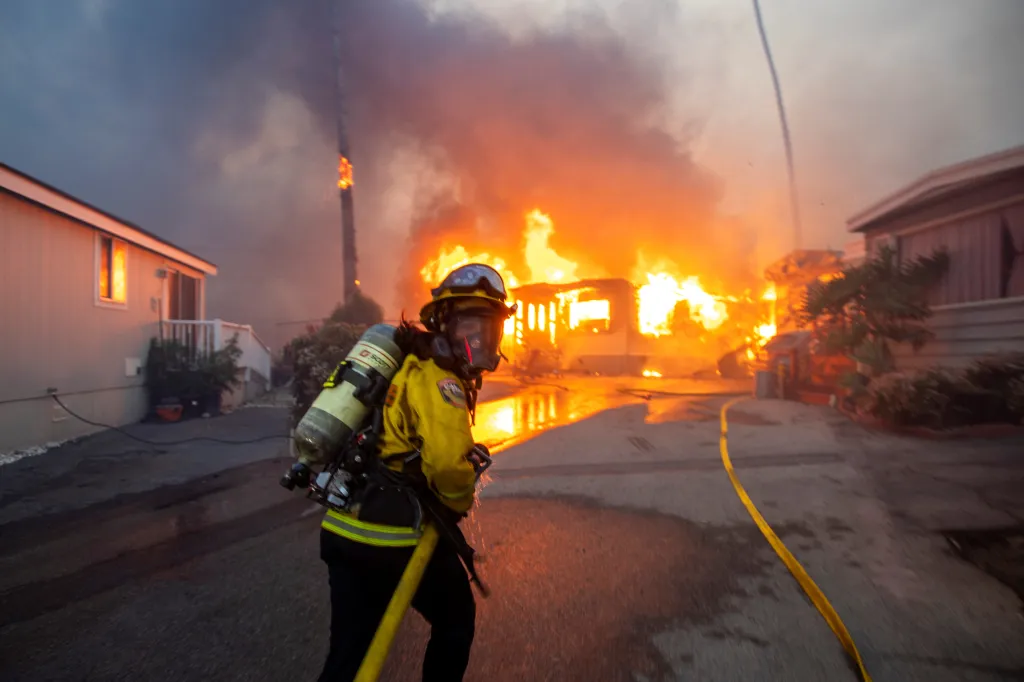 A firefighter in full gear battles the Palisades Fire in Los Angeles, with burning houses and heavy smoke in the background.