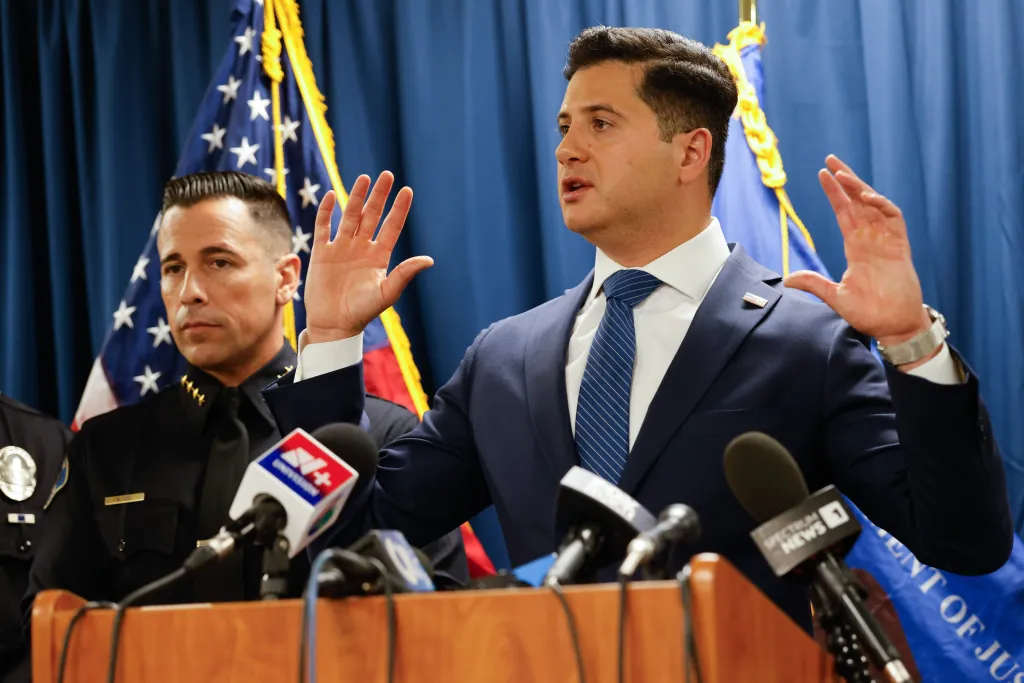 First Assistant United States Attorney Bill Essayli speaking at a press conference at a podium, with a police officer standing next to him and an American flag behind them.