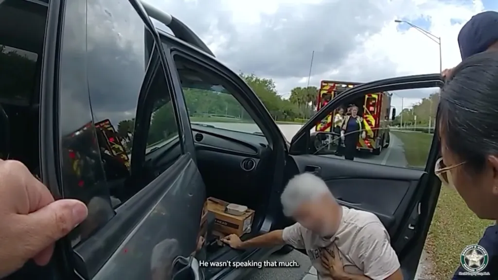 A man holding his chest near an open car door, with an ambulance and first responders visible in the background.