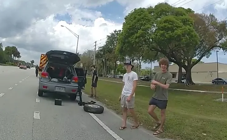 Three teenagers stand by the side of a road next to a disabled car with its trunk open and a flat tire on the ground, with an ambulance in the background.