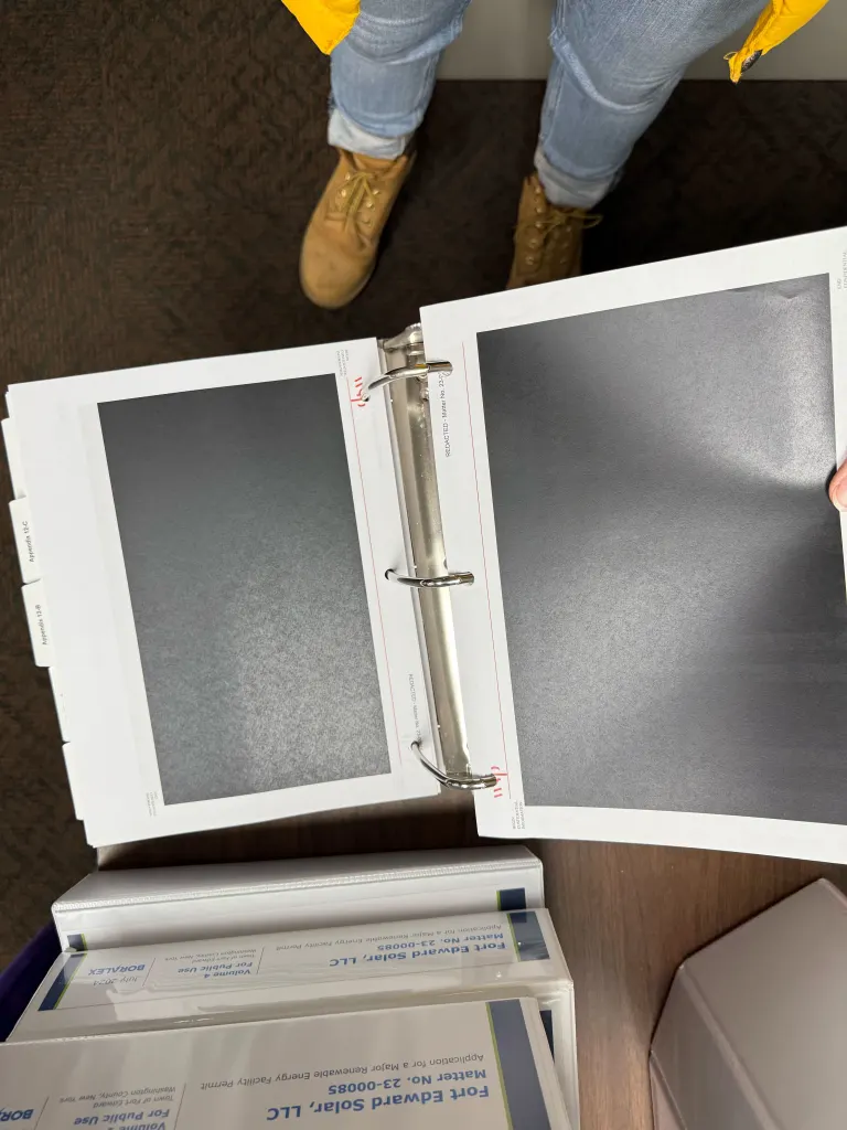 A person holding open a binder filled with pages that have been redacted to hide confidential information regarding the Fort Edward Solar project.