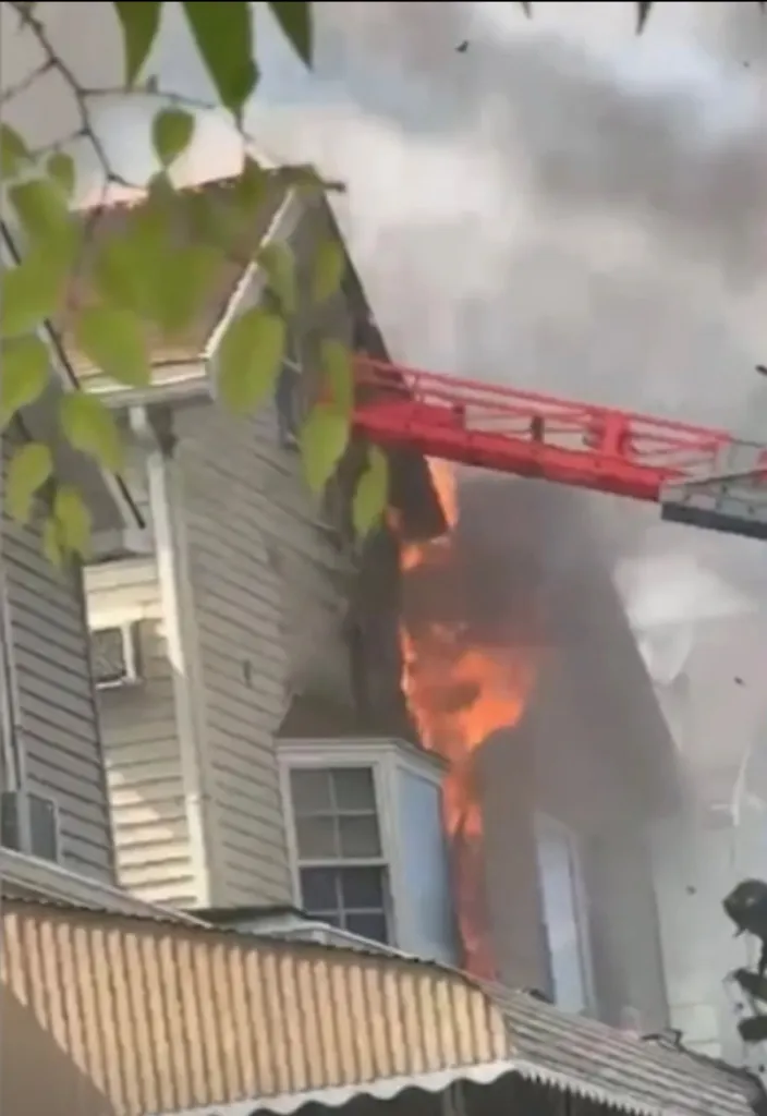 A two-story white house on fire with a red fire truck ladder extended into the top story.