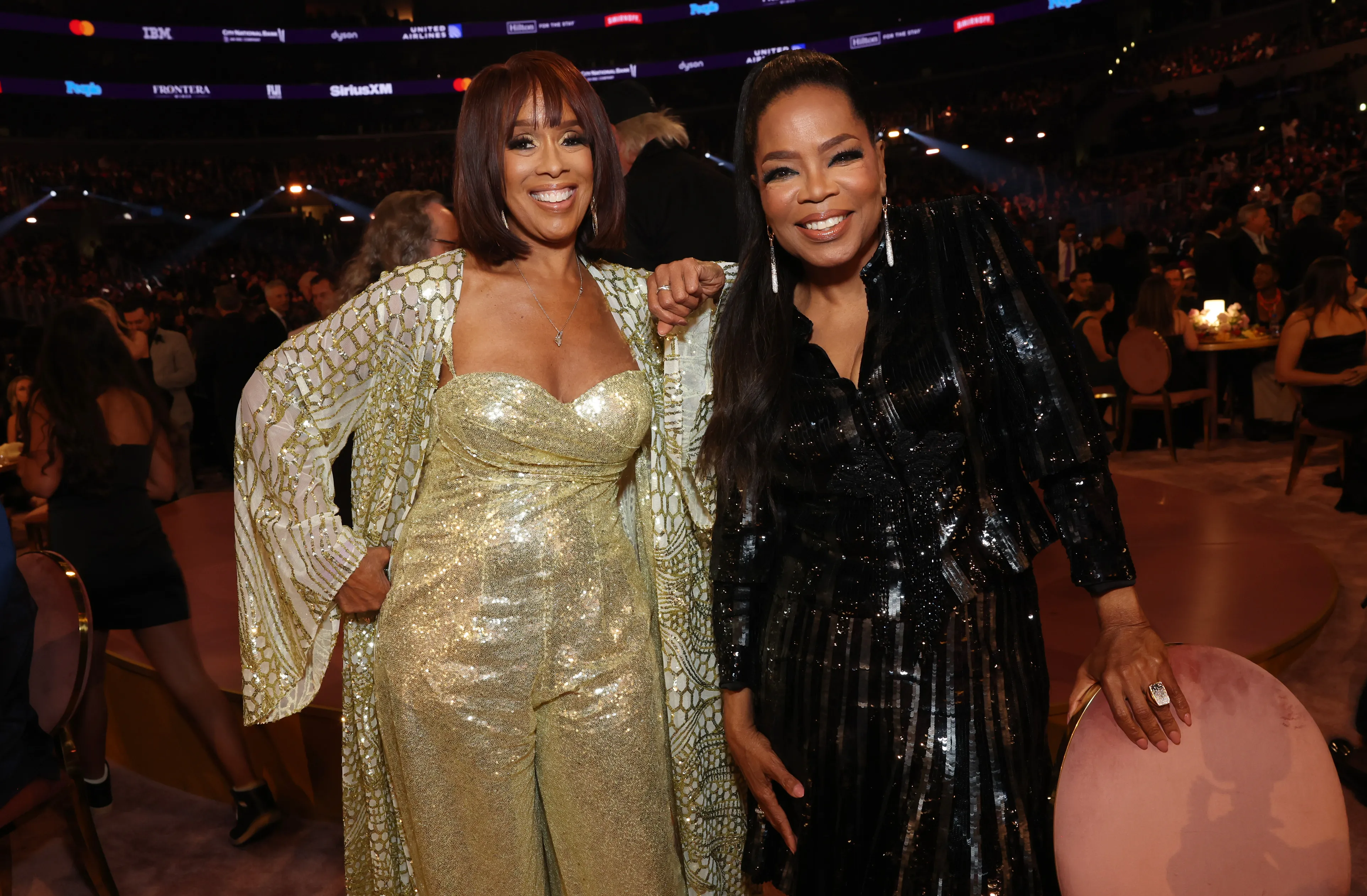 Gayle King in a gold sequined jumpsuit and Oprah Winfrey in a black sequined dress posing together at an event.