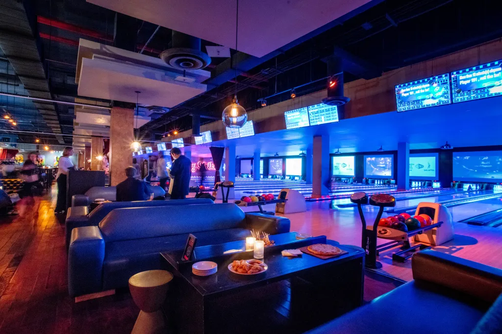 Bowling alley with neon blue lights and several lanes, as well as couches and tables with snacks and drinks.