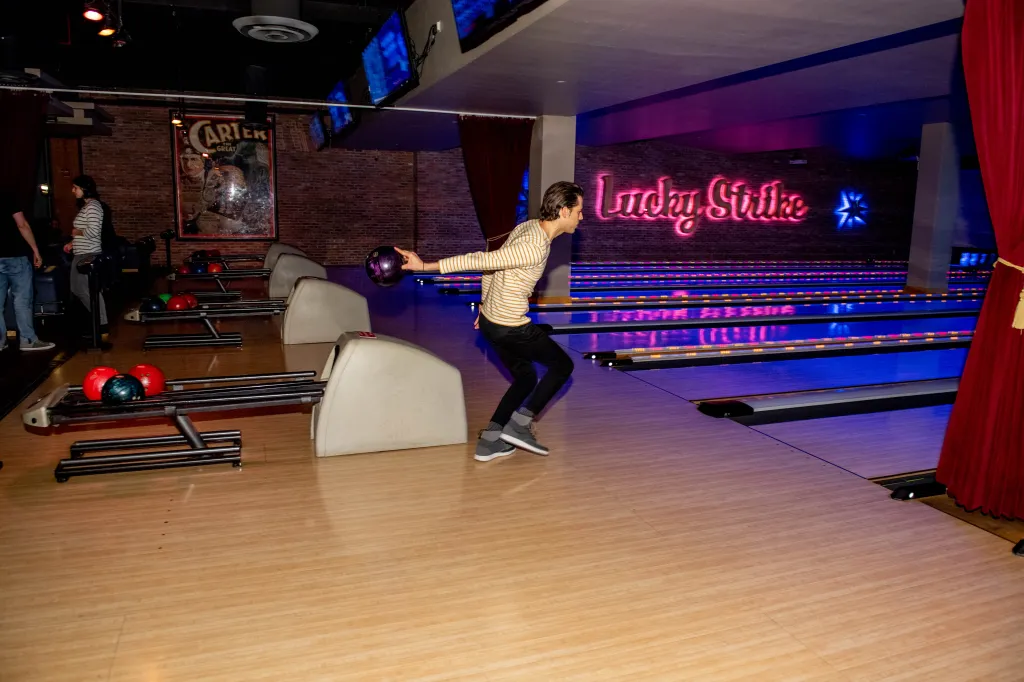 A man in a striped shirt and dark pants bowling in a dim bowling alley with blue and pink lights.