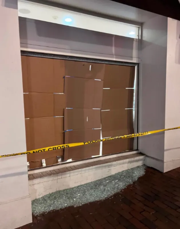 A shattered storefront window, boarded up with cardboard and surrounded by police tape and broken glass.