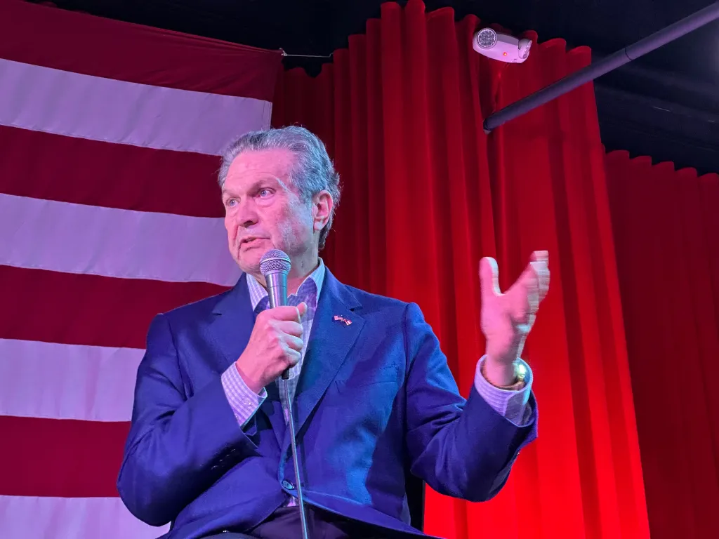 Rick Jackson speaking with a microphone in front of an American flag and red curtains.