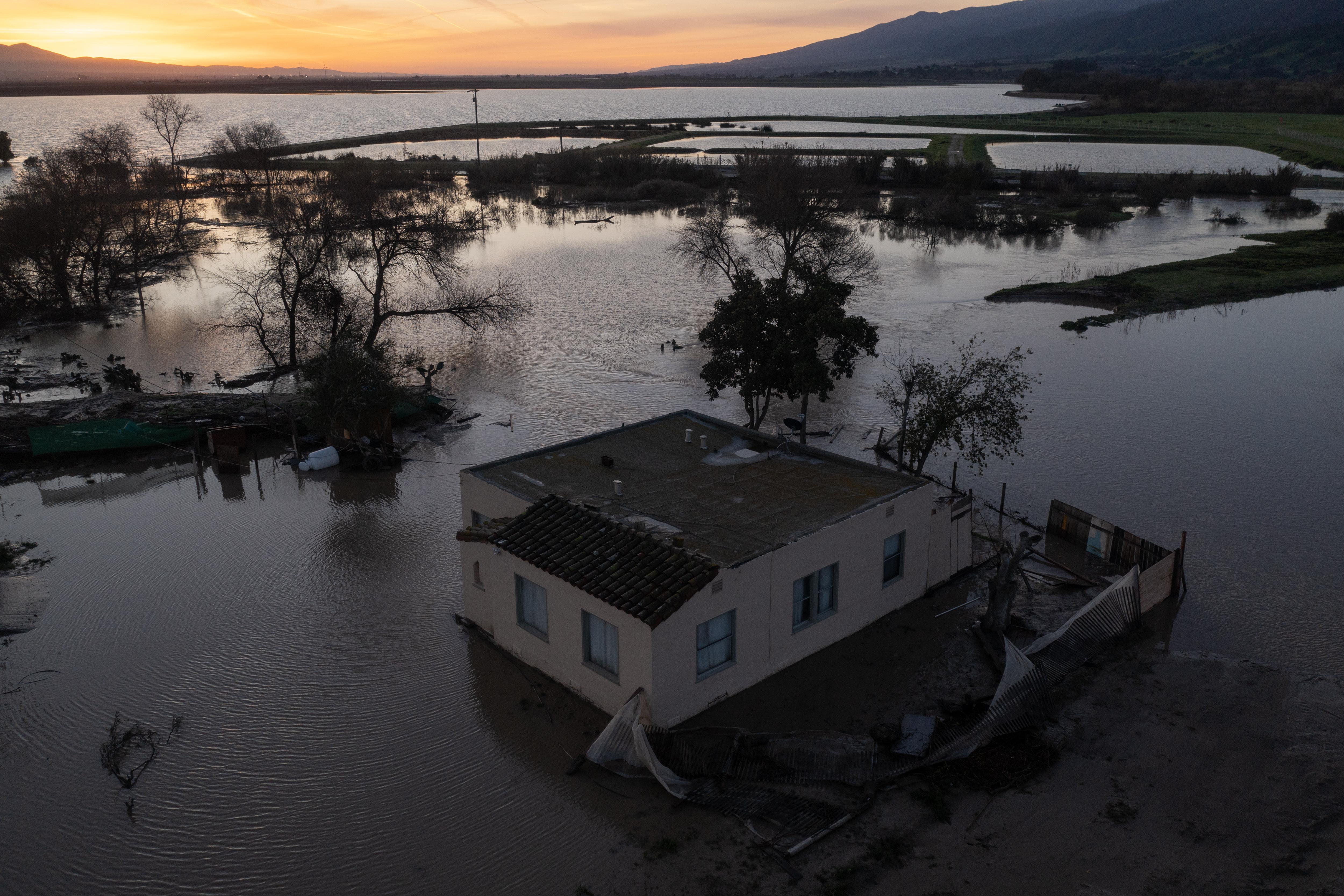a house shown surrounded by flood waters and partially submerged trees with mountains in the background 