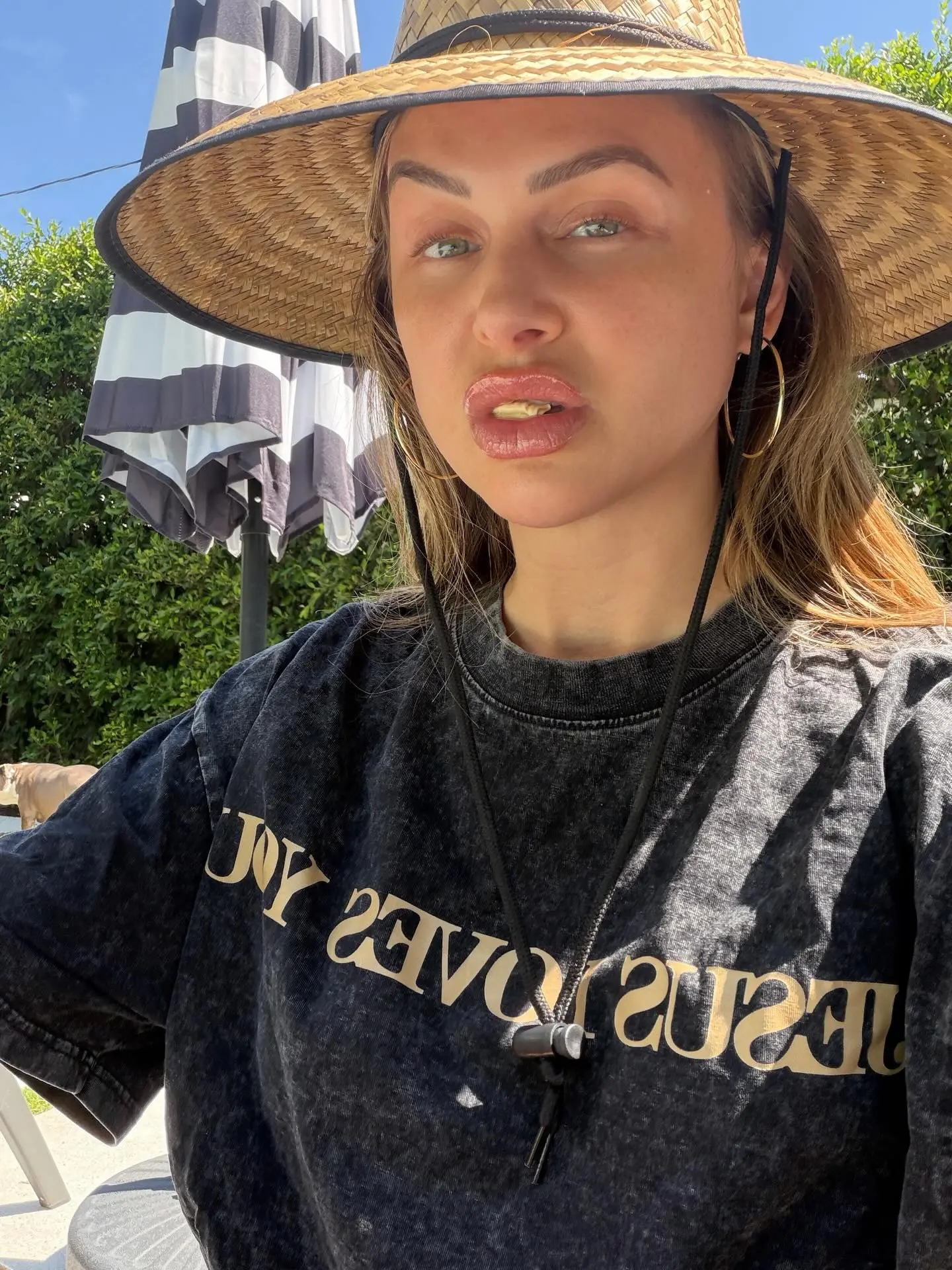 Lala Kent in a straw hat and black t-shirt, with a partial view of a striped umbrella and green foliage in the background.