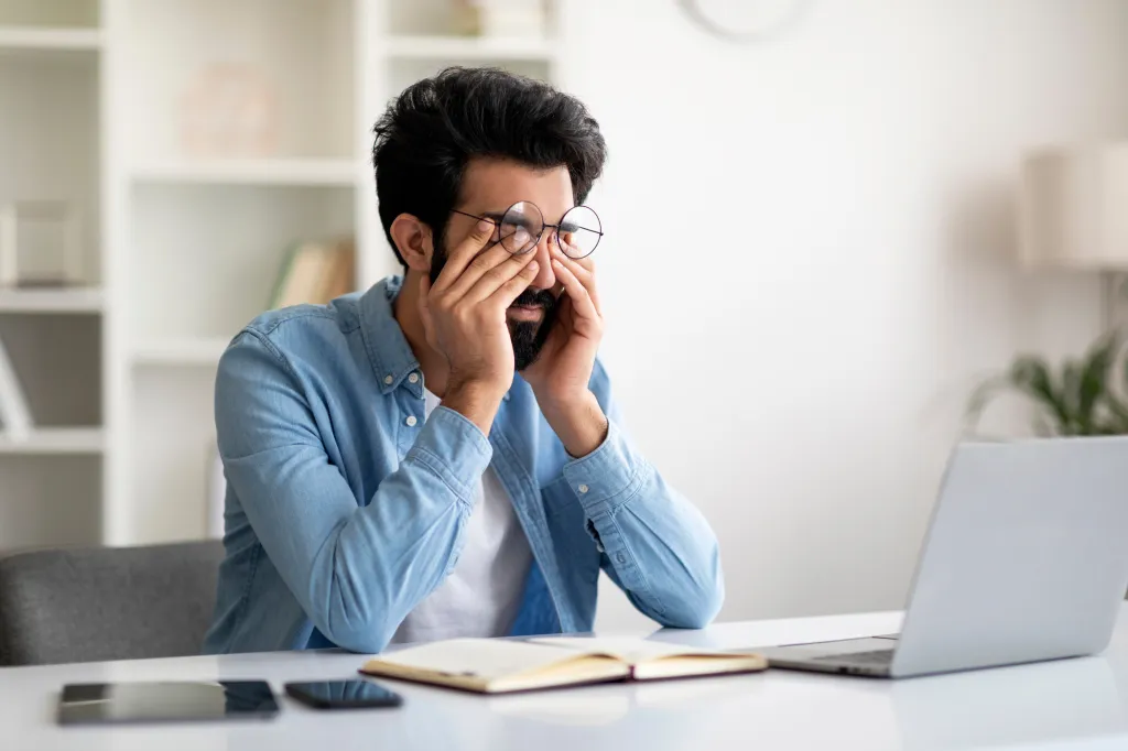 Young Indian man with round glasses massaging his tired eyes after working on a laptop.