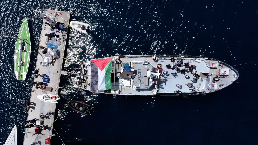 Aerial view of the Global Sumud Flotilla's main vessel with a Palestinian flag, loaded with people and supplies, docked alongside a smaller boat and a pier.