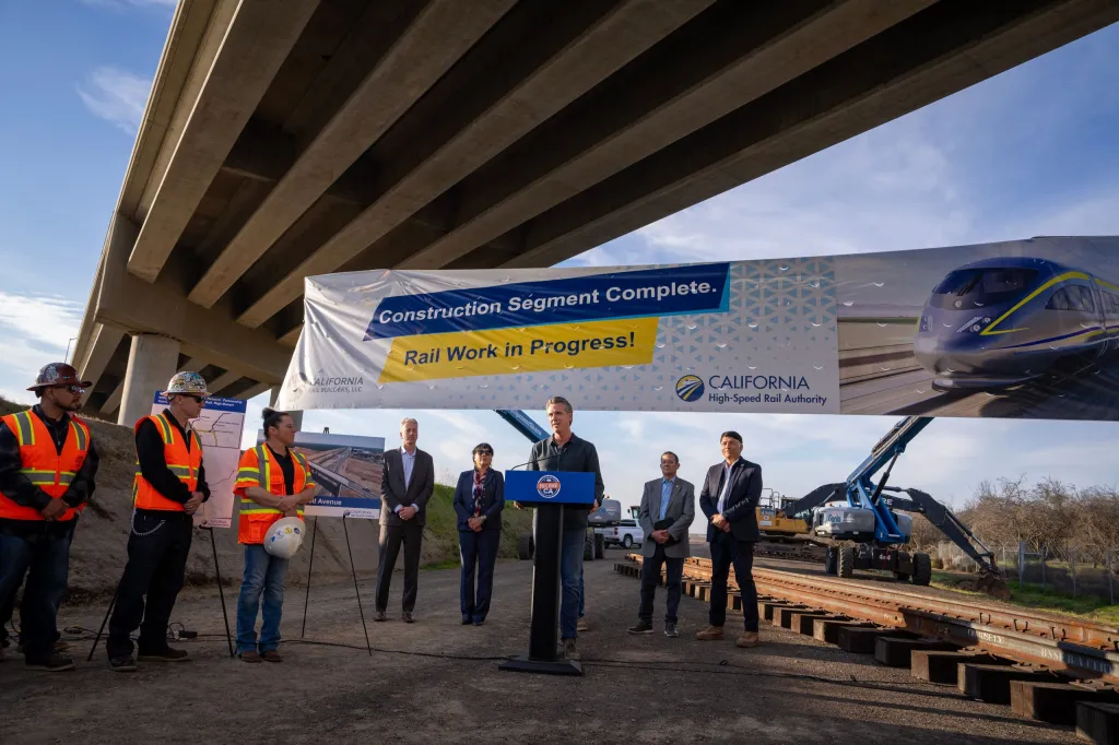 Governor Gavin Newsom speaking at a podium under an overpass, next to a large banner about the California High-Speed Rail project.