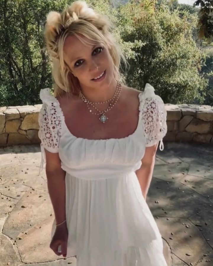 Britney Spears in a white dress and layered necklaces smiling outdoors.