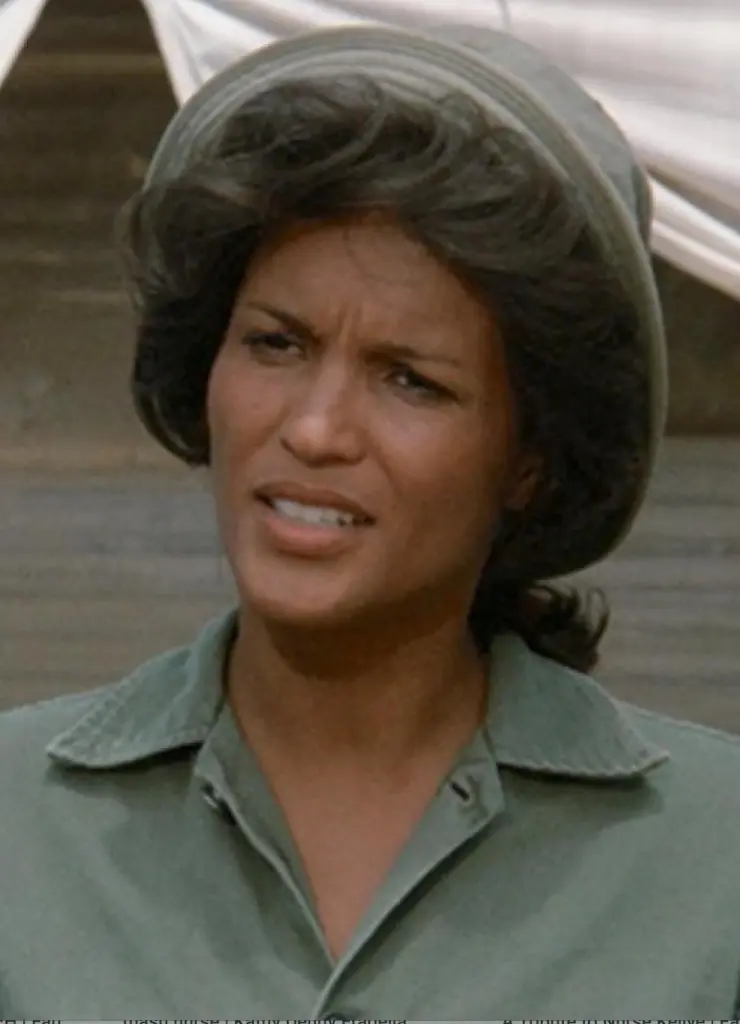 Gwen Farrell in M*A*S*H, wearing a green military-style shirt and cap, with a look of concern on her face.