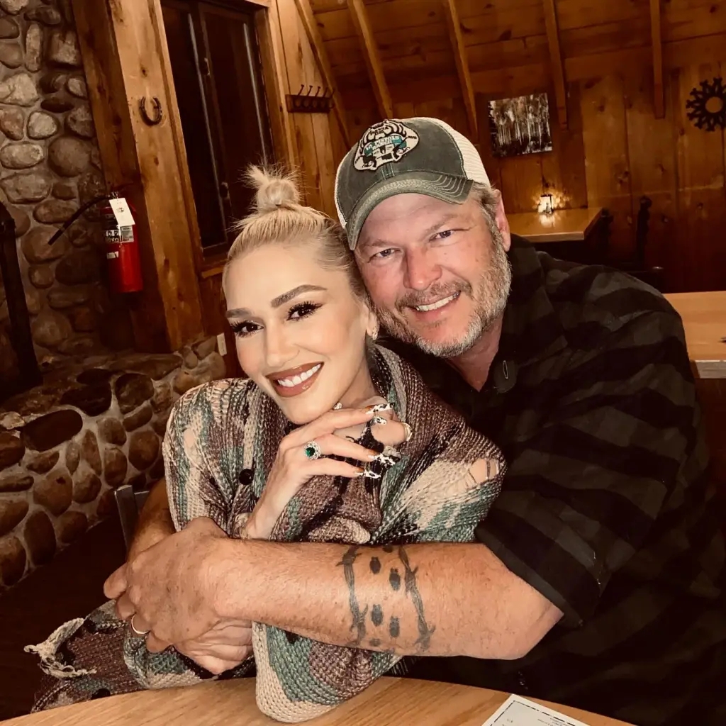 Gwen Stefani and Blake Shelton smiling while embracing at a wooden table in a rustic restaurant.